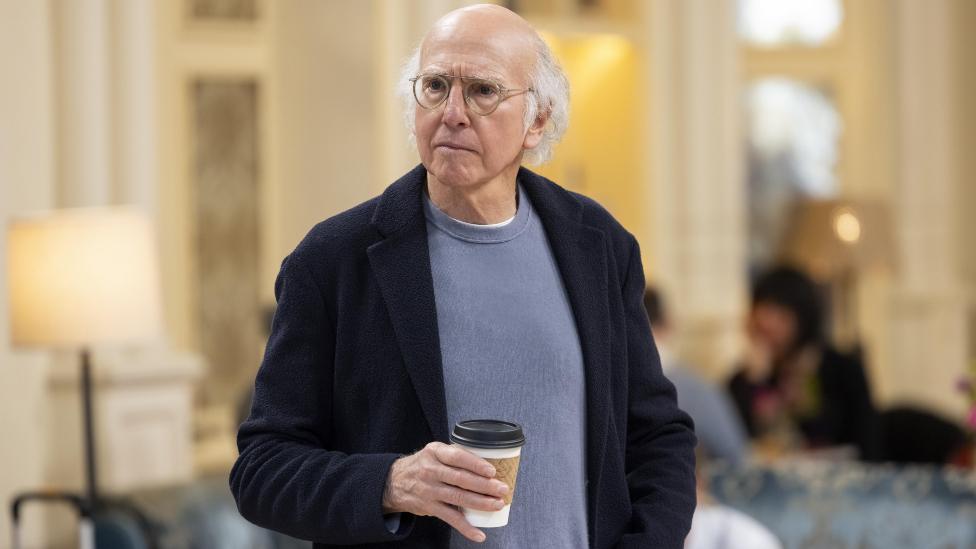 Larry David holding a coffee cup on the set of Curb Your Enthusiasm, dressed casually and looking thoughtful.