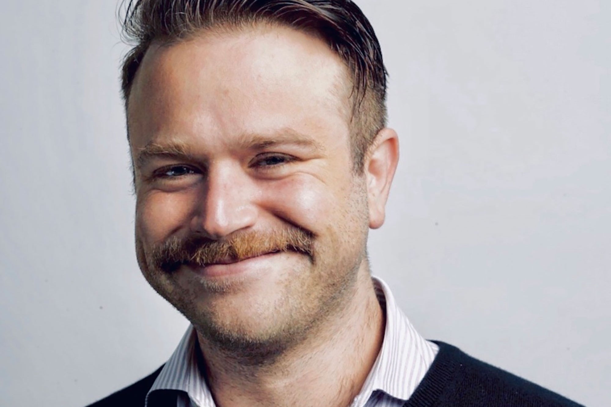 Close-up portrait of Zak Williams smiling warmly, wearing a striped collared shirt under a dark sweater, with neatly styled hair and a mustache.