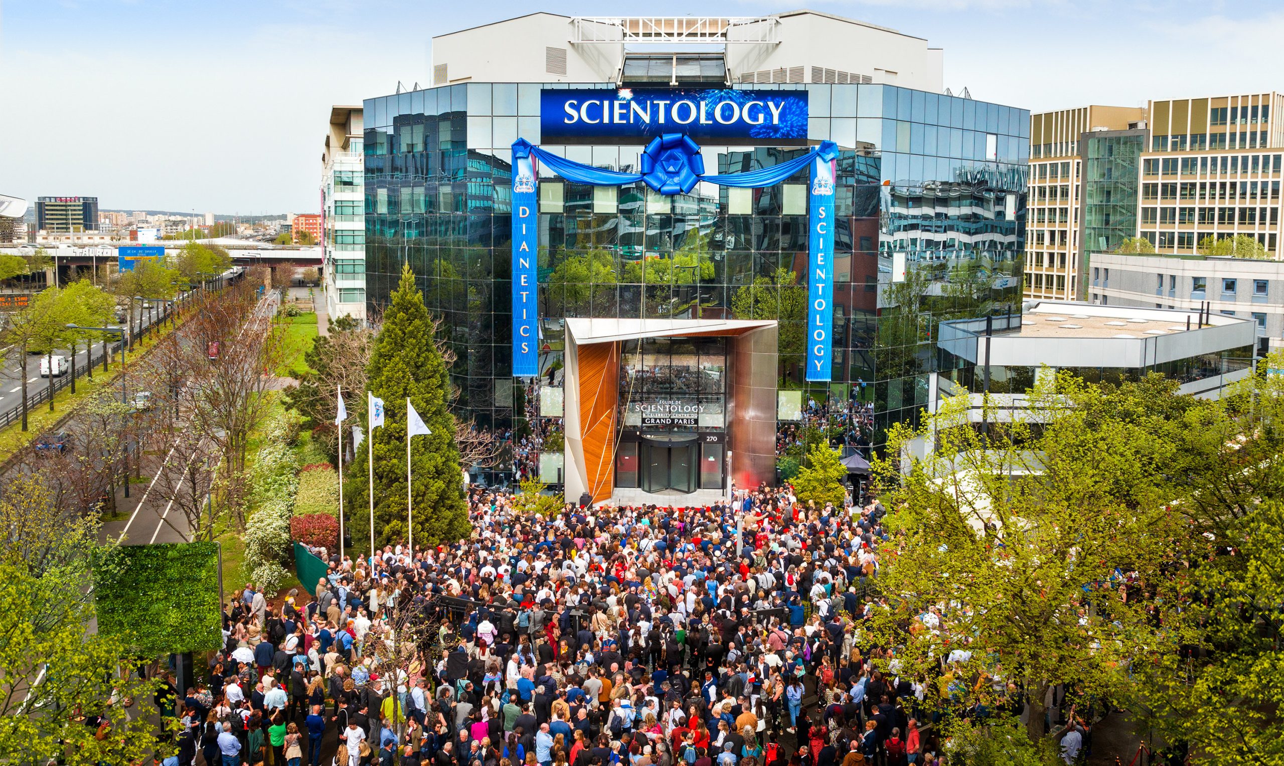 Large crowd gathered outside the modern glass-fronted Church of Scientology Grand Paris building during a grand opening event, with blue banners reading “Dianetics” and “Scientology” and lush greenery surrounding the scene.