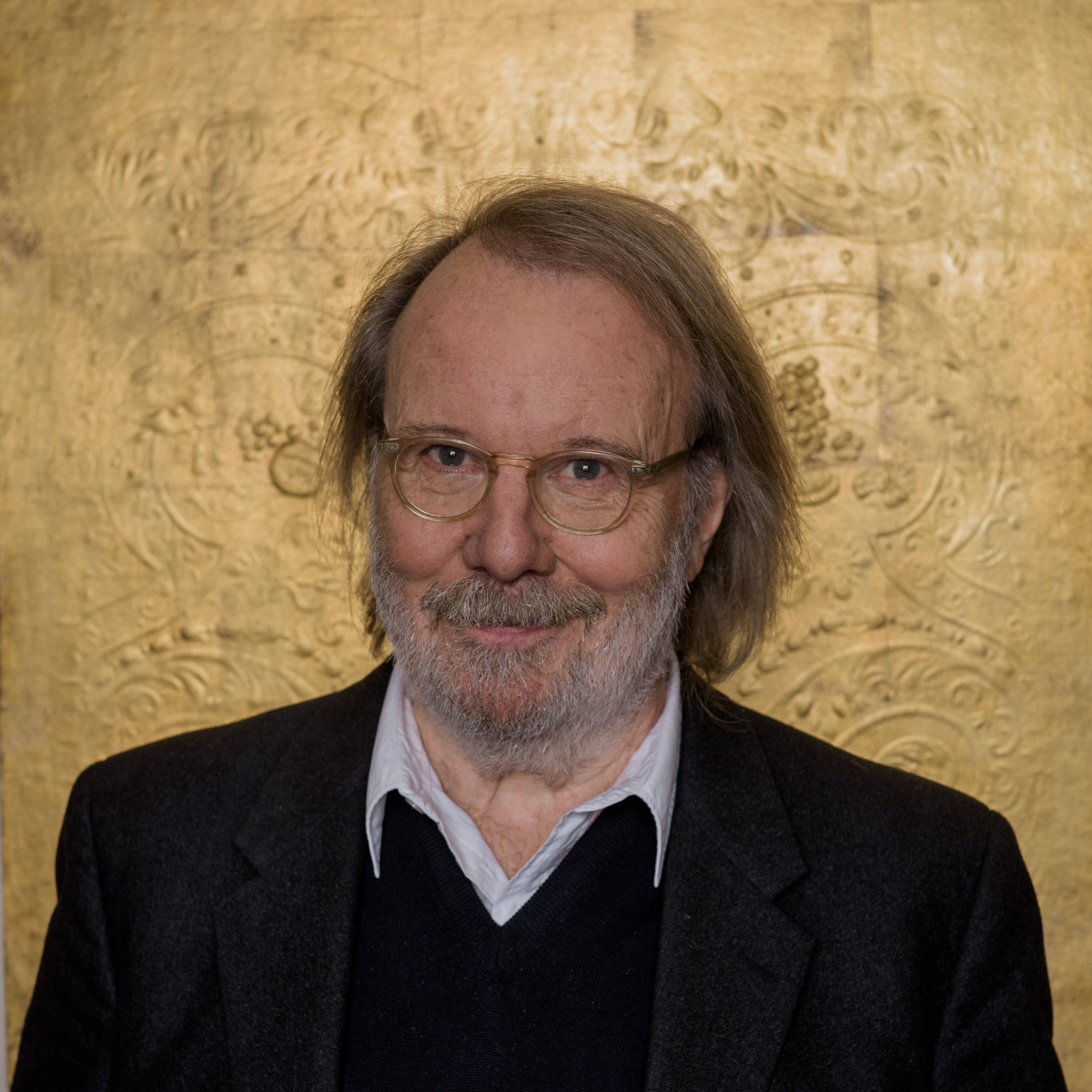 Benny Andersson smiling warmly at the camera with a shimmering gold background.