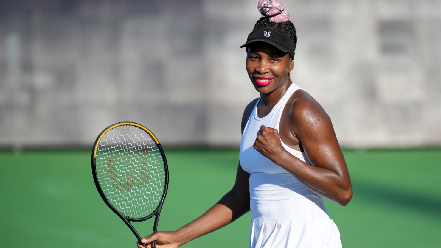 Venus Williams smiling warmly at the camera while standing on the grass courts of Wimbledon, dressed in her classic tennis whites.