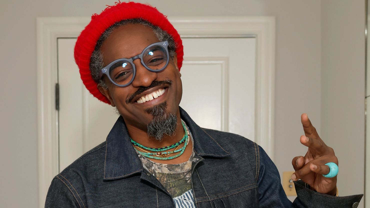 André 3000 smiling warmly at the camera, wearing a stylish hat and patterned shirt, exuding relaxed confidence.