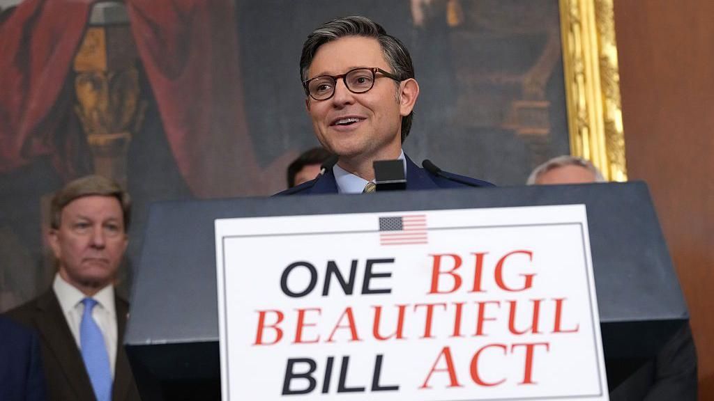 A smiling man in a suit and glasses speaks at a podium with a sign reading "ONE BIG BEAUTIFUL BILL ACT" in bold letters, with an American flag icon above the text. Other suited individuals stand behind him in a formal room with classic artwork and gold-framed decor.