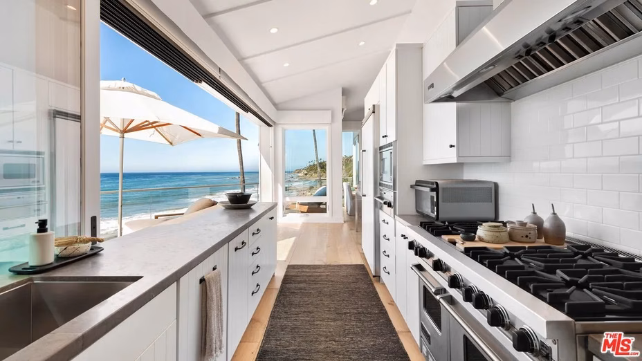 Sleek, narrow kitchen with minimalist cabinetry and premium appliances, seamlessly opening into a sunlit living room with dramatic ocean views.