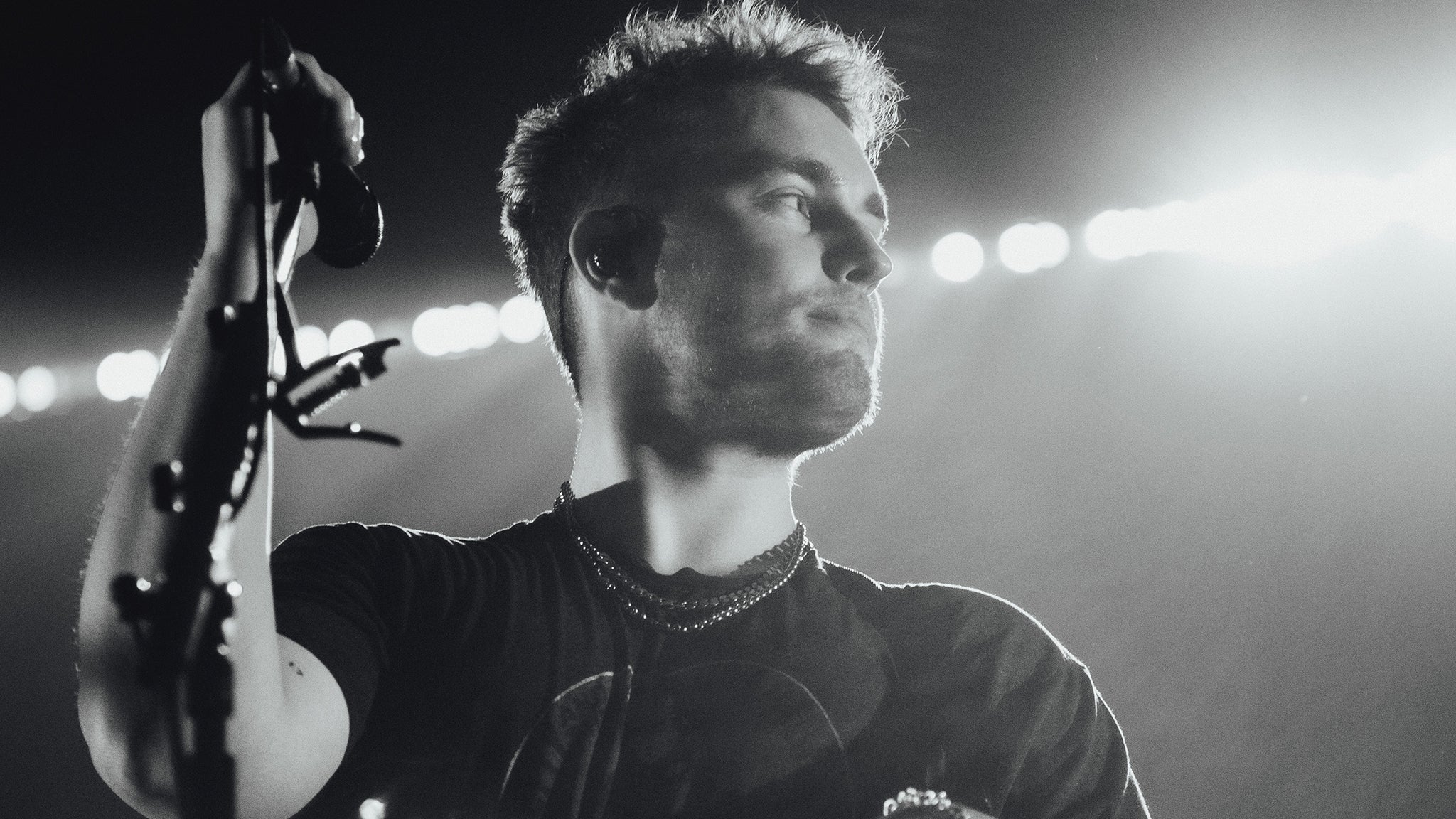 Sam Fender performing live in black and white, with dramatic stage lights illuminating him from behind.