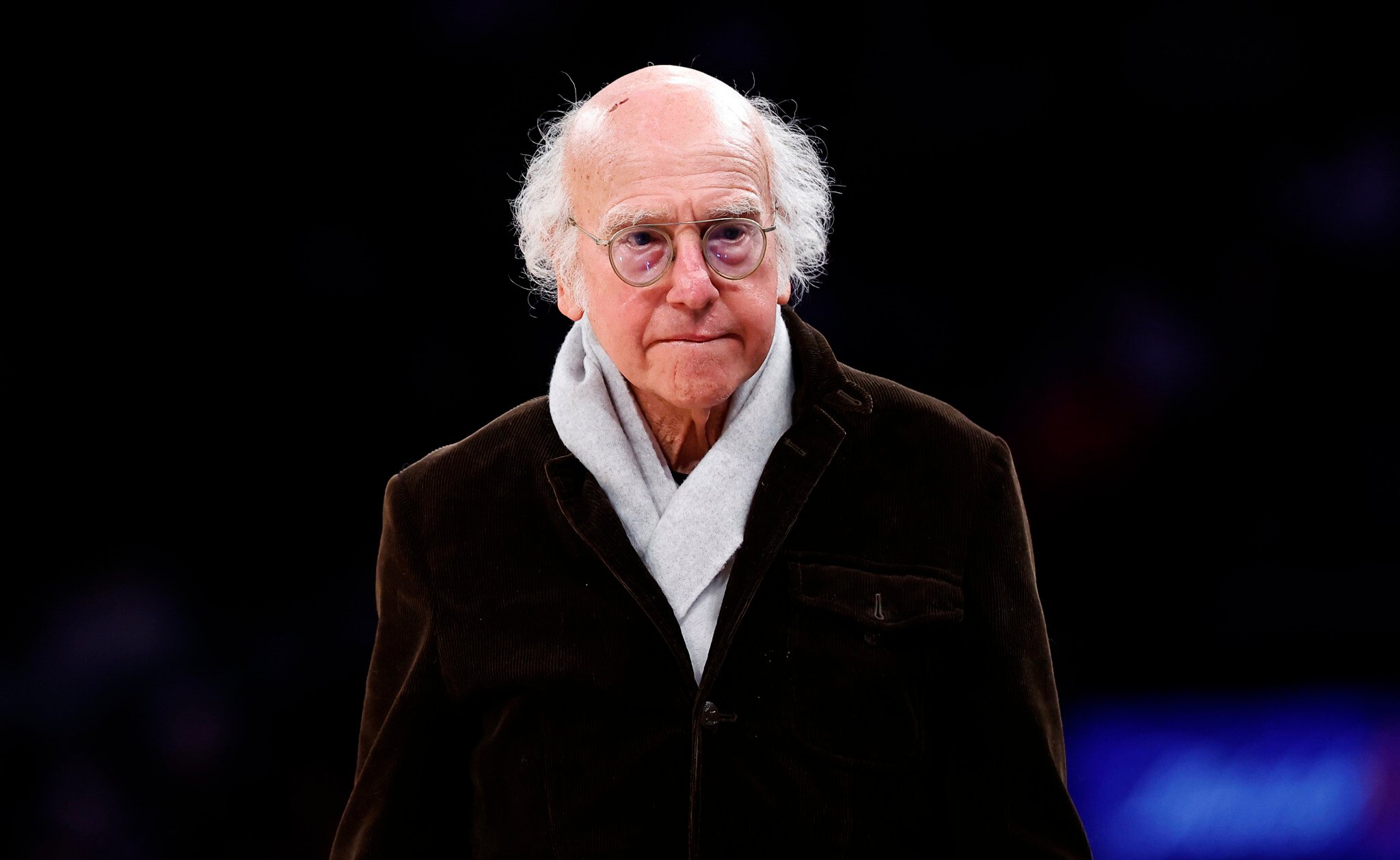 Larry David walking outdoors wearing a coat and scarf on a cool day.