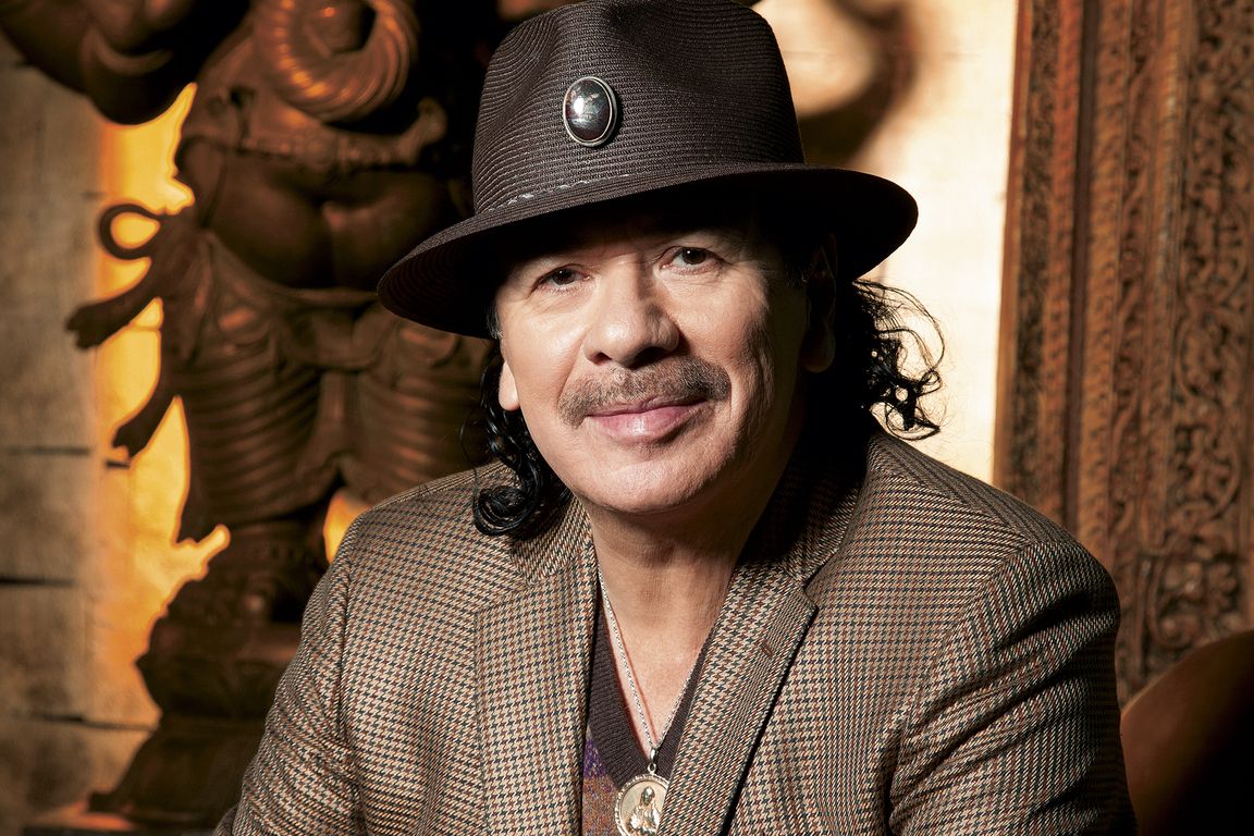 Carlos Santana smiling warmly during a professional photo shoot, dressed casually with soft lighting.