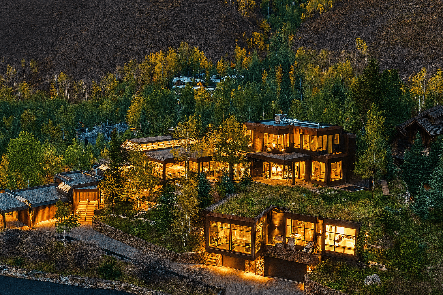 Aerial view of James Hetfield's modern luxury mountain home in Vail, Colorado, surrounded by trees and hills, illuminated warmly at dusk.