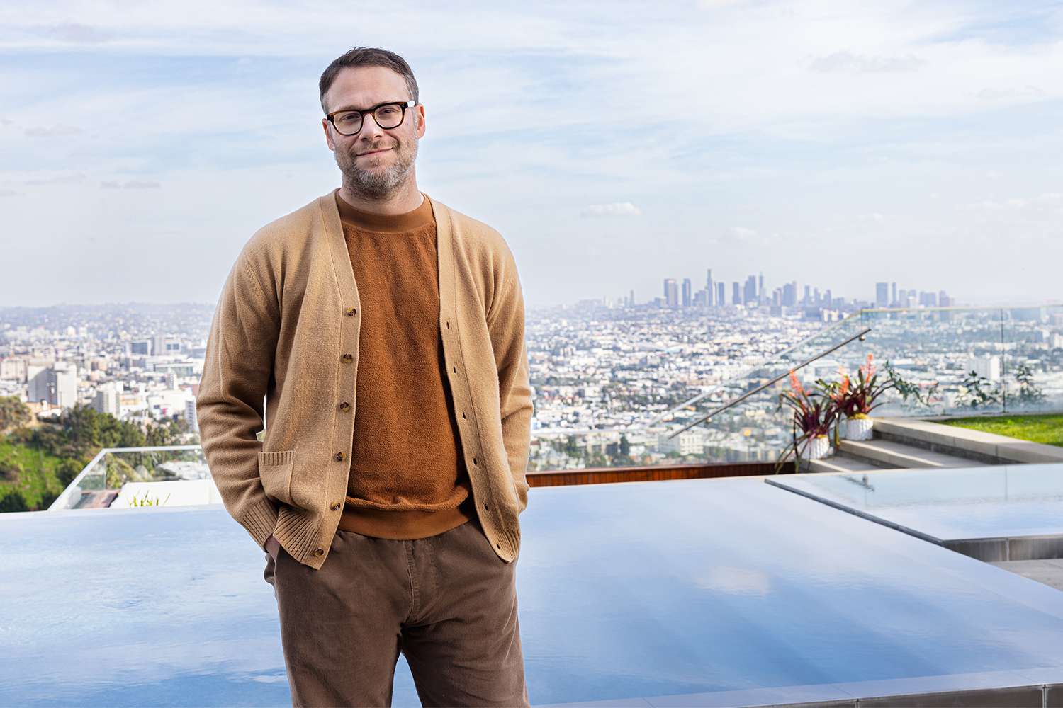 Seth Rogen smiling by a swimming pool with a panoramic view of the Los Angeles skyline behind him.