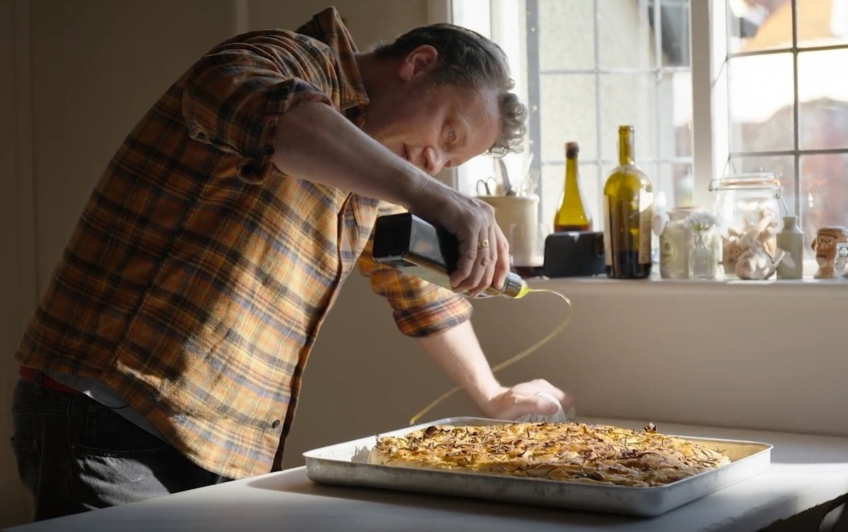 Jamie Oliver drizzling olive oil over freshly baked focaccia bread in a rustic kitchen setting.