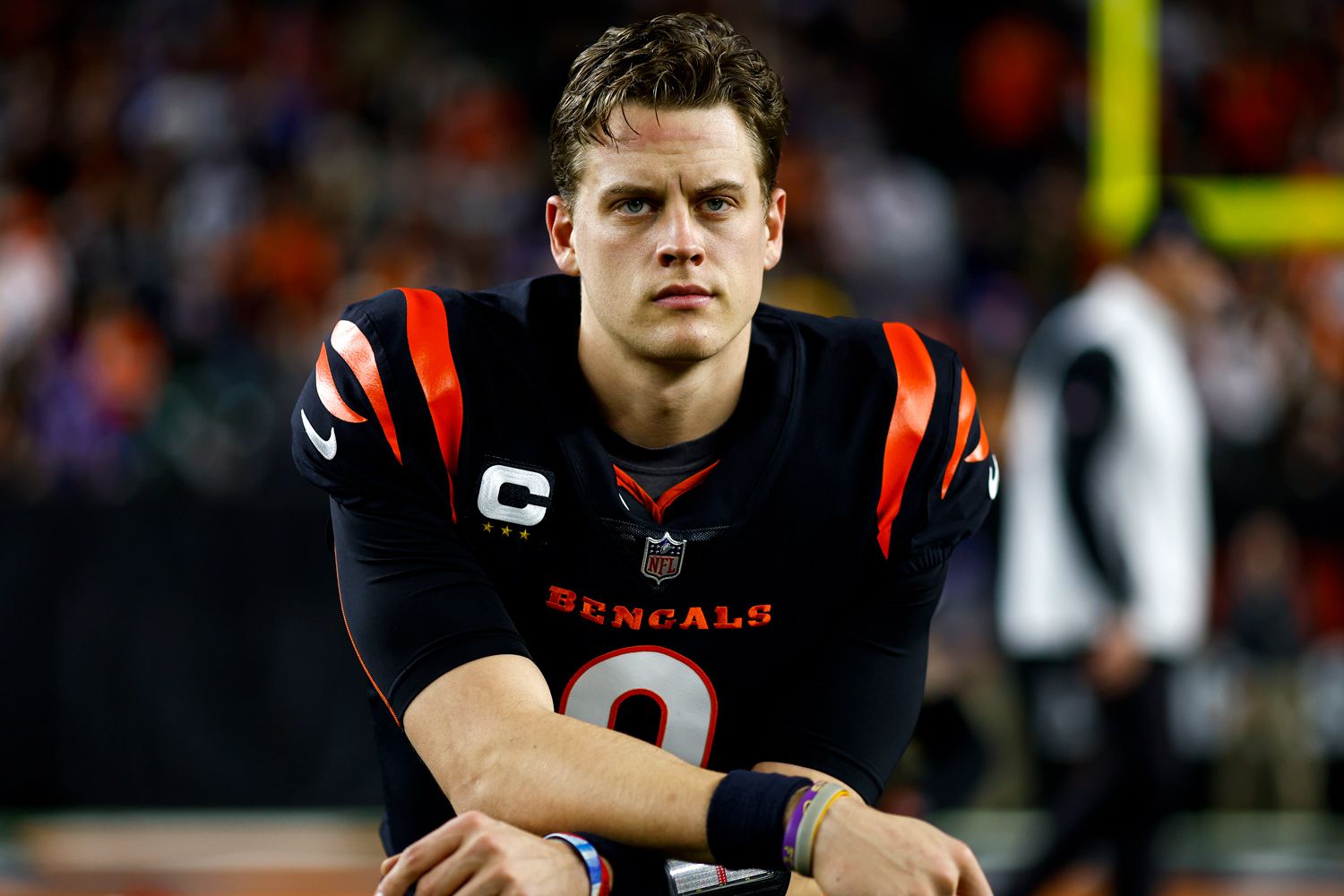 NFL quarterback in a black Cincinnati Bengals uniform with orange stripes, kneeling on the field with a serious expression. The player wears a captain’s patch, wristbands, and the number 9 on his jersey.