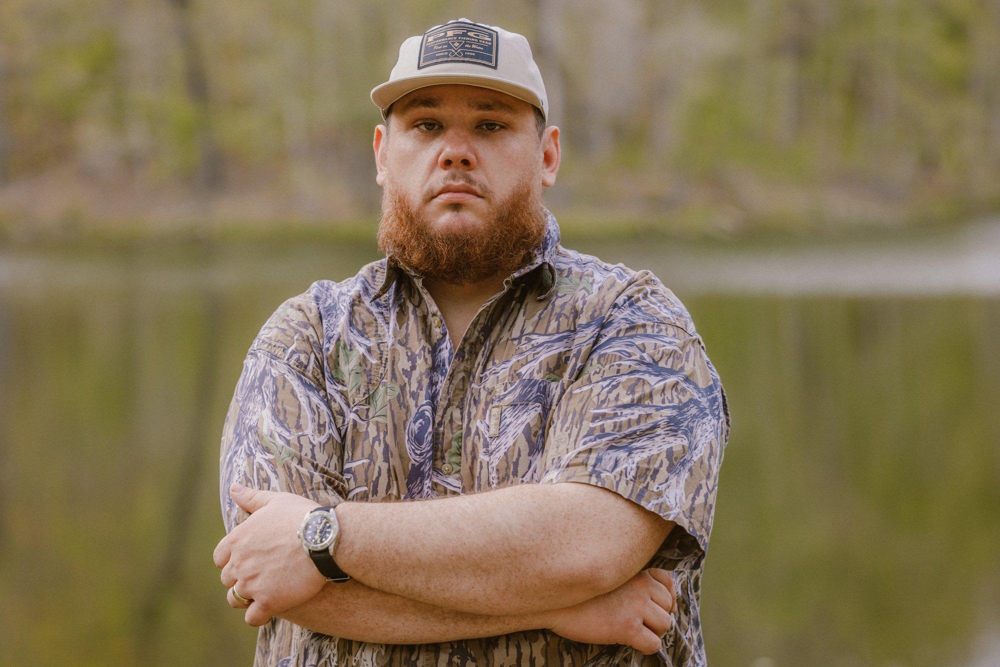 Luke Combs posing outdoors in a camouflage shirt, smiling at the camera with natural scenery in the background.