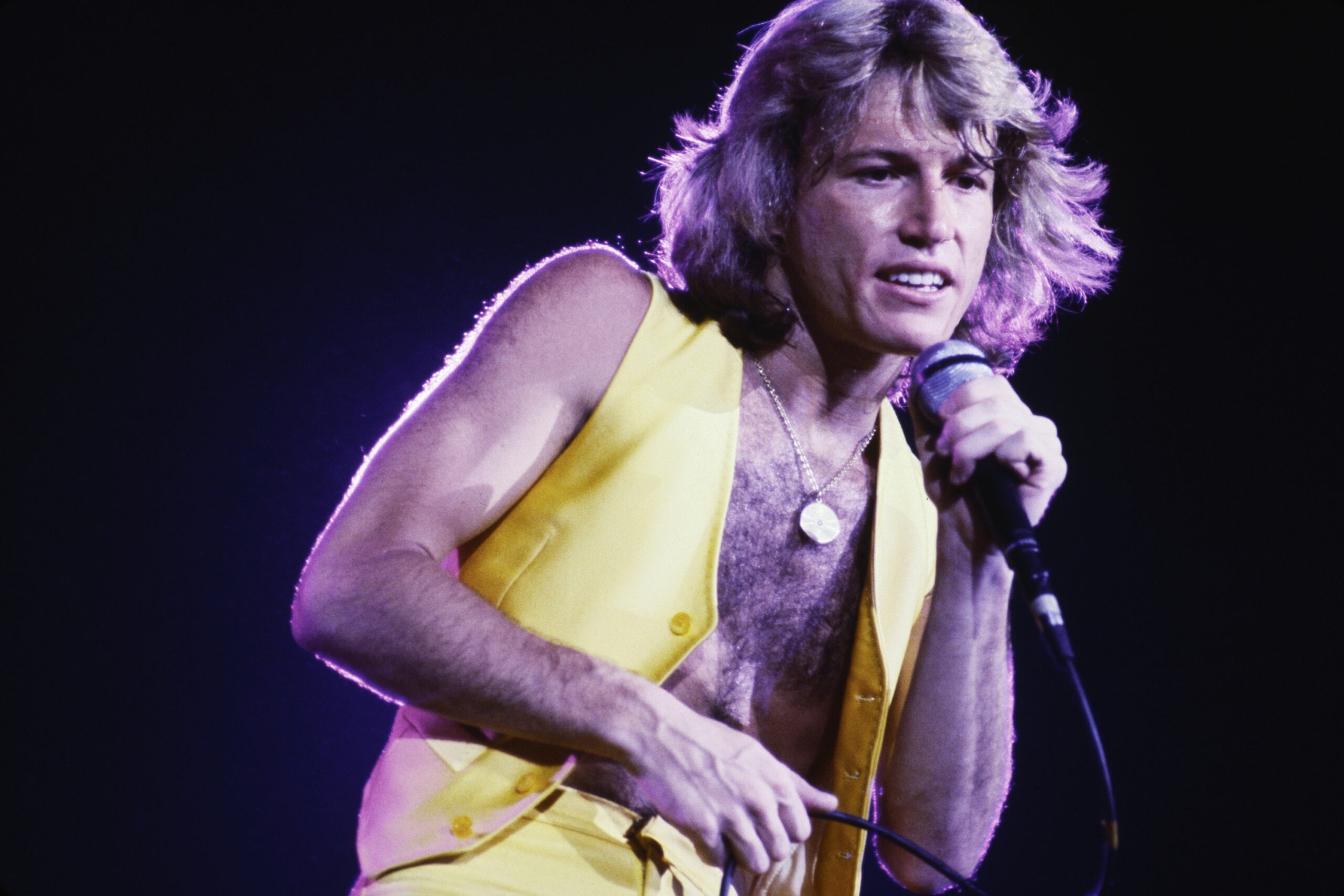 Andy Gibb performing on stage wearing a bright yellow waistcoat, mid-song under stage lights