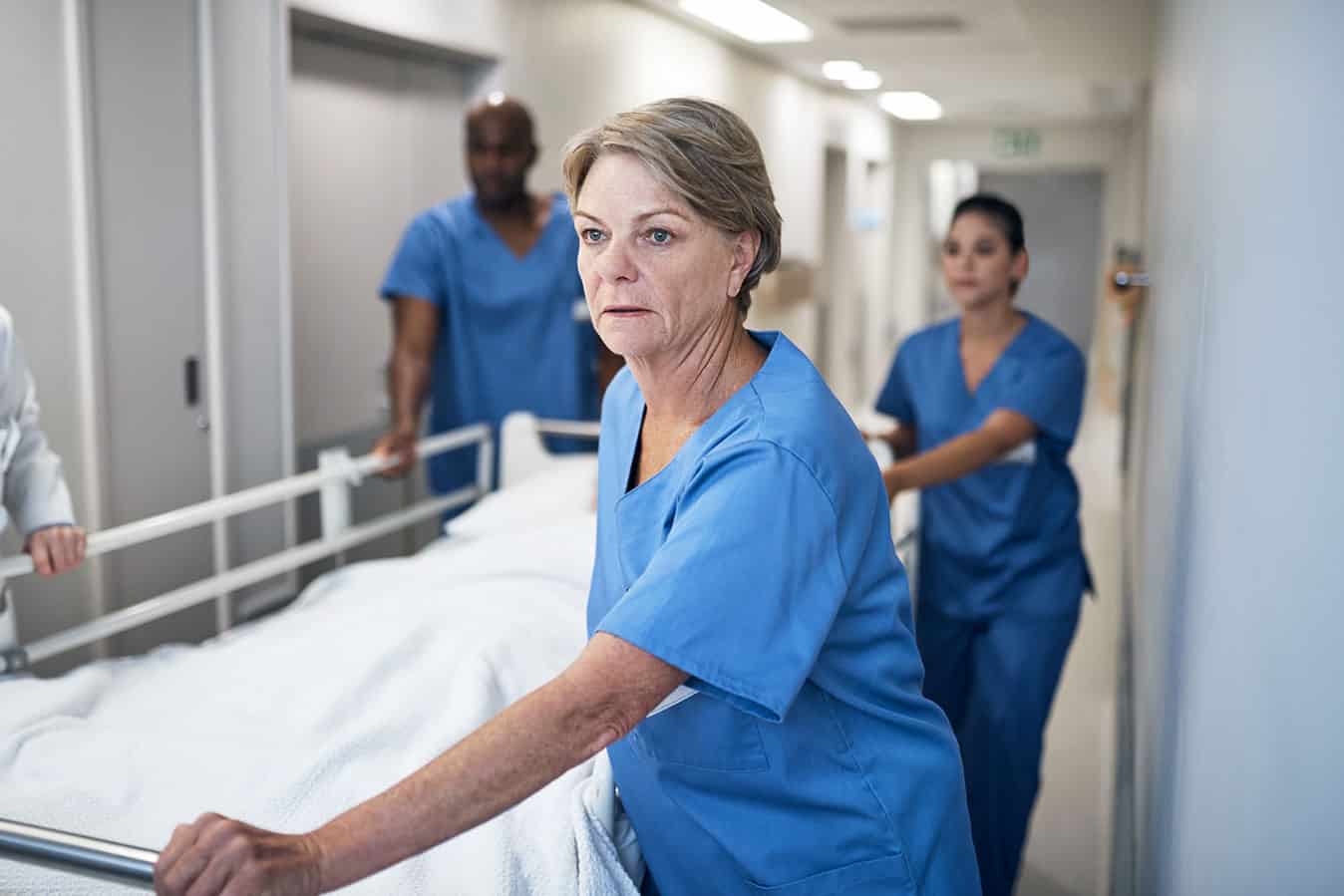 Older female nurse pushing hospital bed in hallway, looking tired, with healthcare team in background.