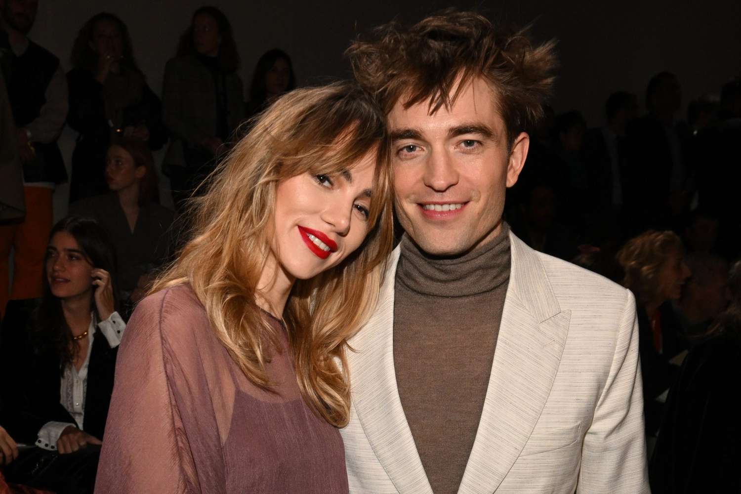 Robert Pattinson and Suki Waterhouse posing together on the red carpet, both elegantly dressed and smiling.