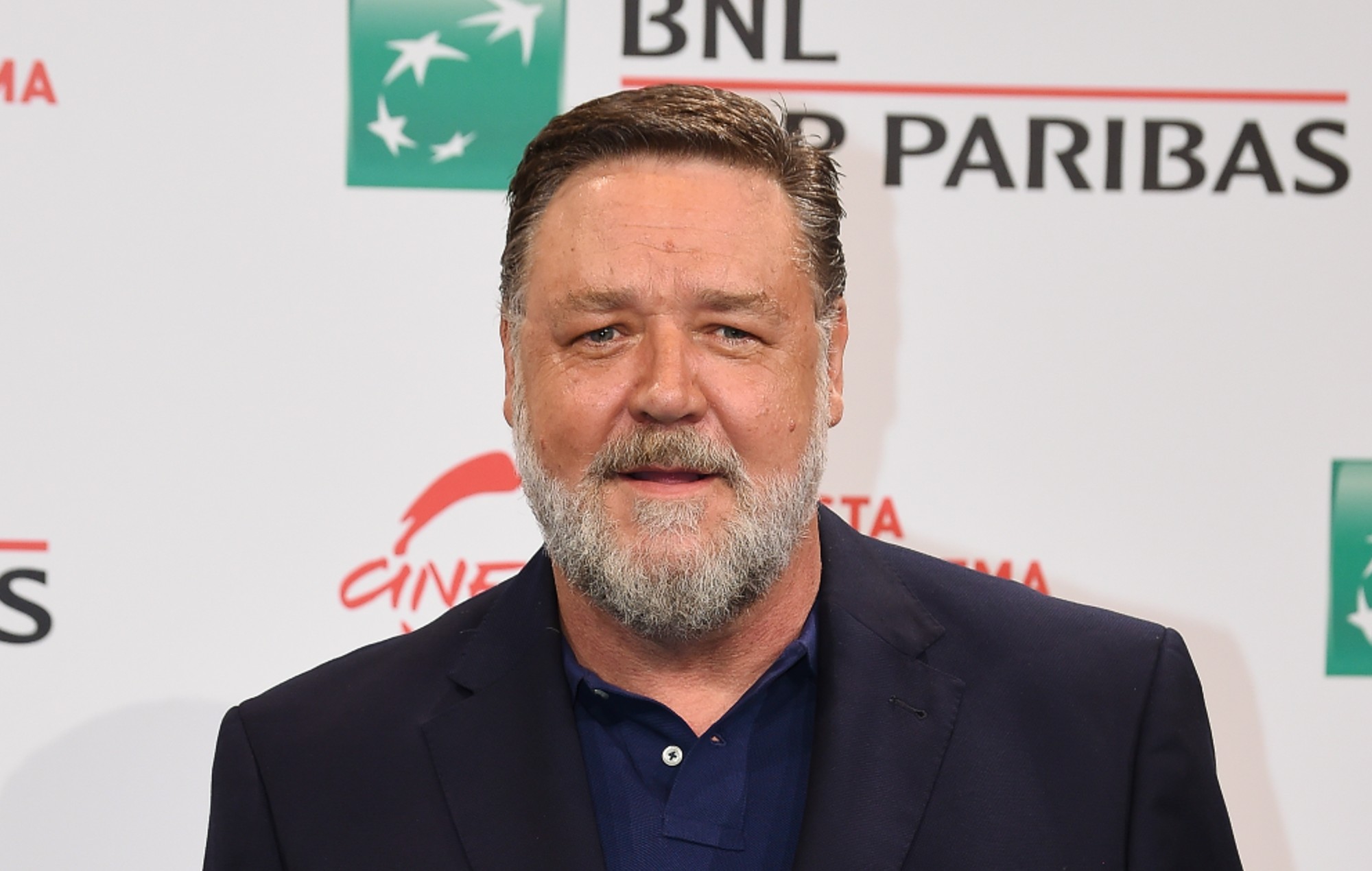 Russell Crowe smiling confidently at the camera while posing on the red carpet in a dark suit.