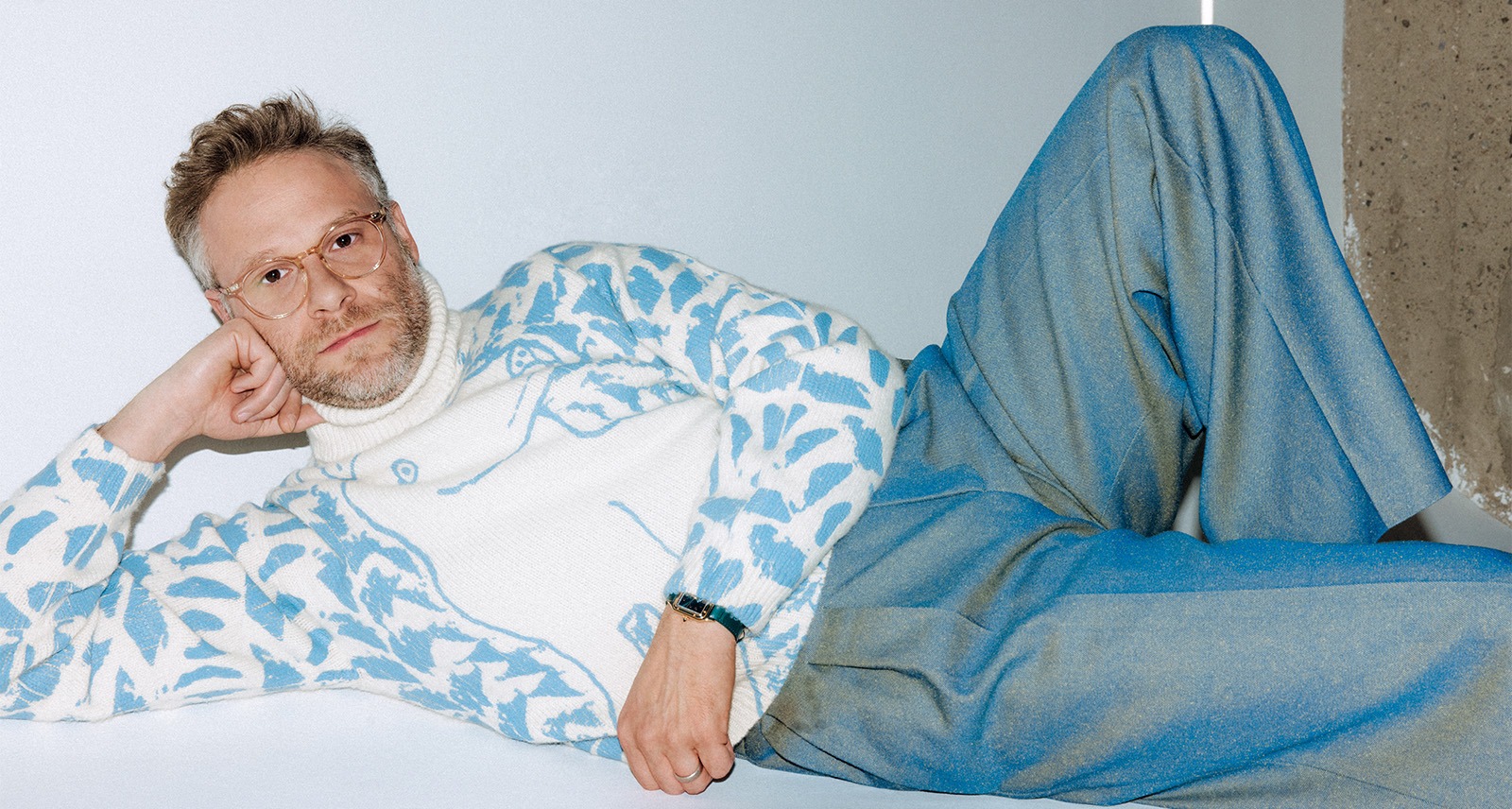 Seth Rogen lying down on the floor, striking a playful and humorous pose with a big smile.
