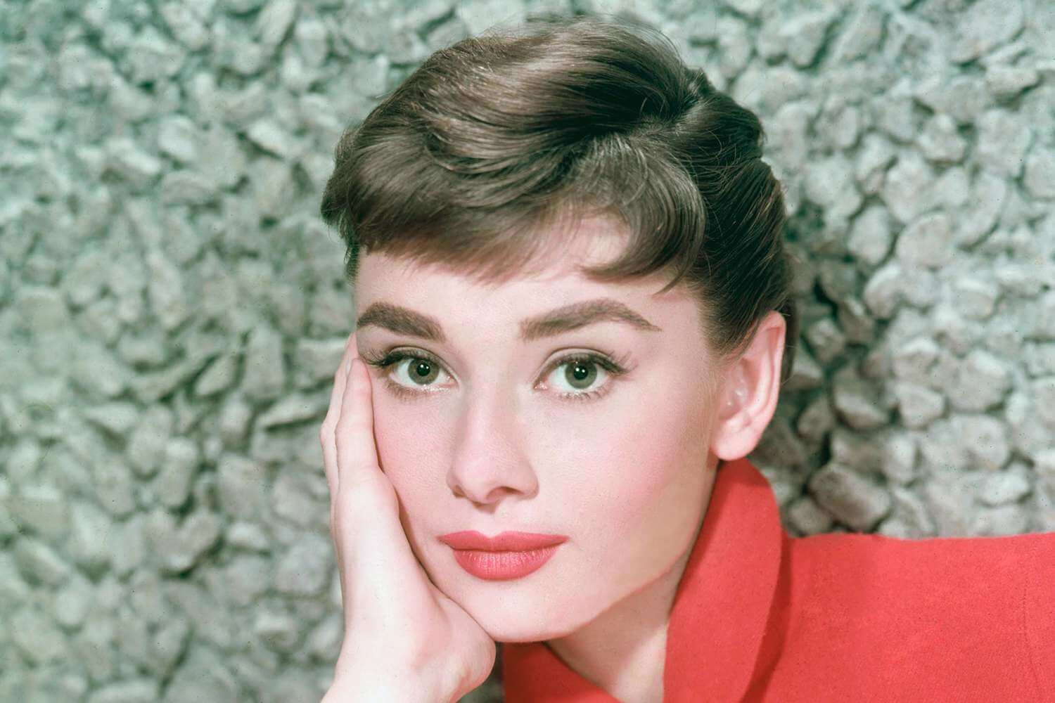 Audrey Hepburn posing confidently in a red shirt with matching red lipstick, looking directly at the camera.