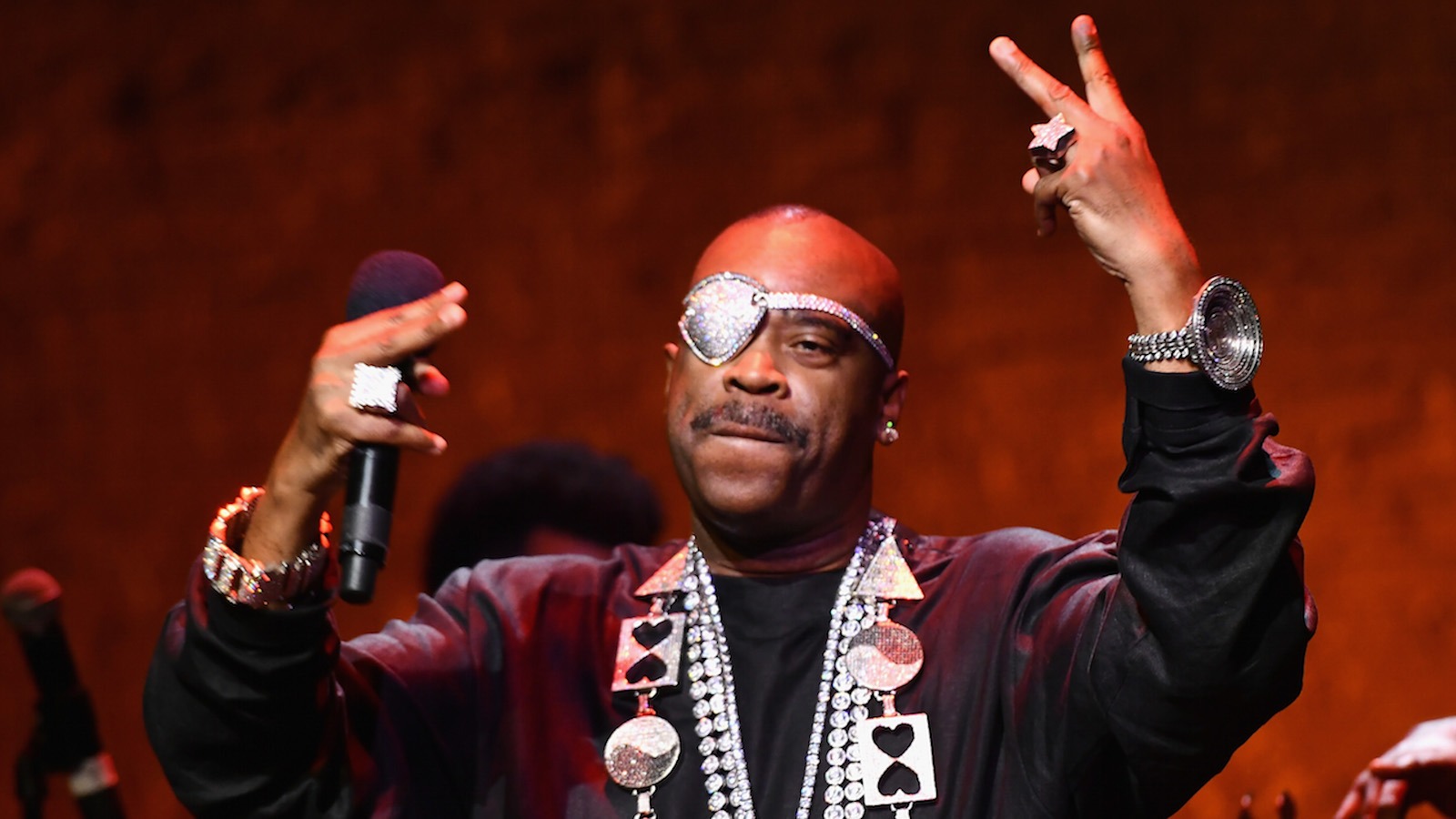 Slick Rick throwing up gang signs toward the crowd during a live performance, dressed in bold streetwear and signature jewelry.