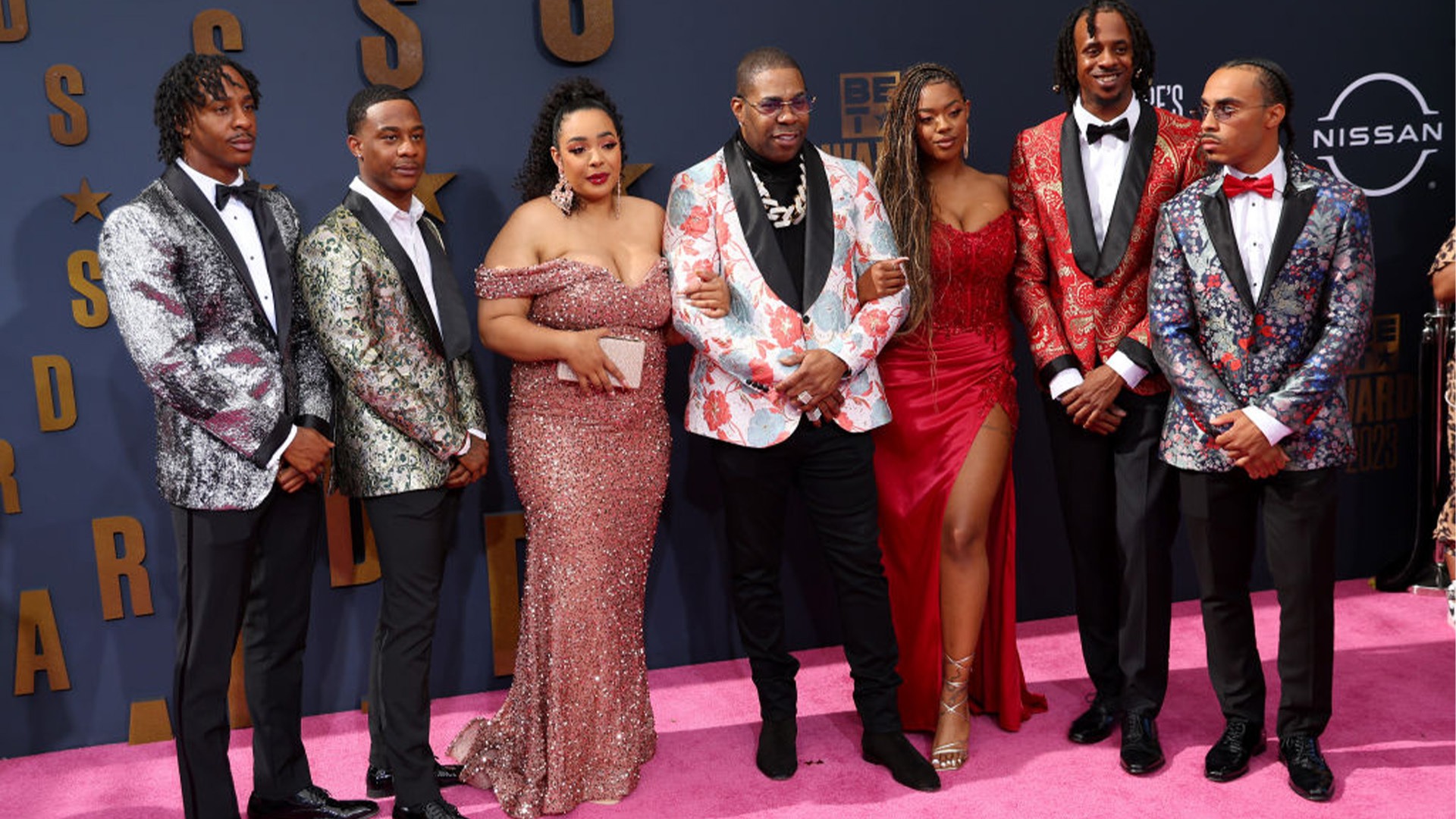 Busta Rhymes standing proudly with his six children on the red carpet, all dressed in coordinated formal attire.