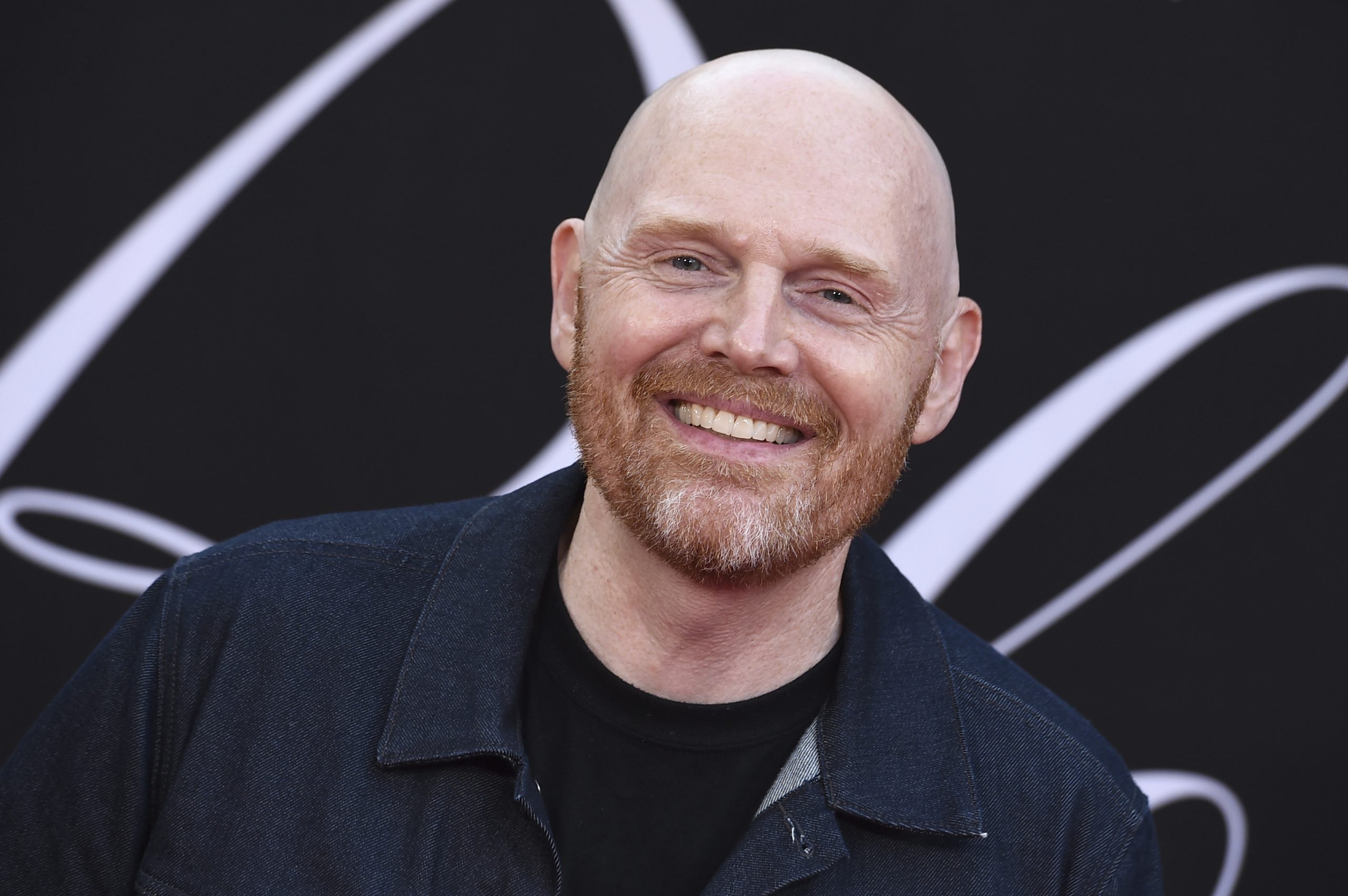 Bill Burr smiling on the red carpet, wearing a dark jacket and black shirt, against a black background with white design elements.