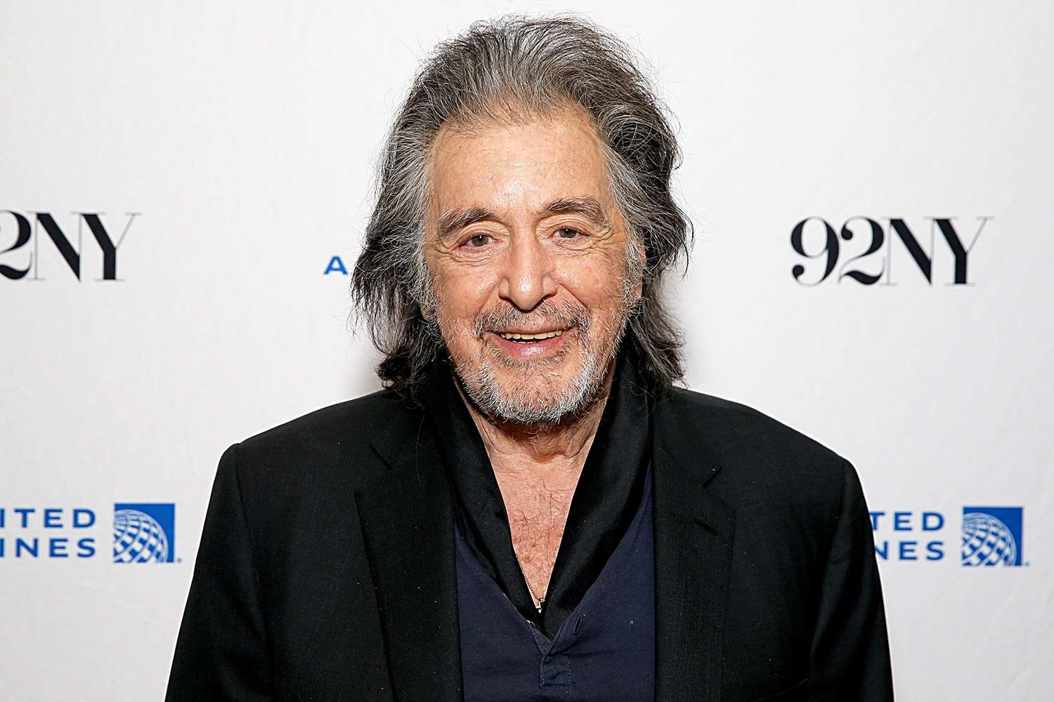 Al Pacino smiles at a public event, wearing a black jacket against a white backdrop.