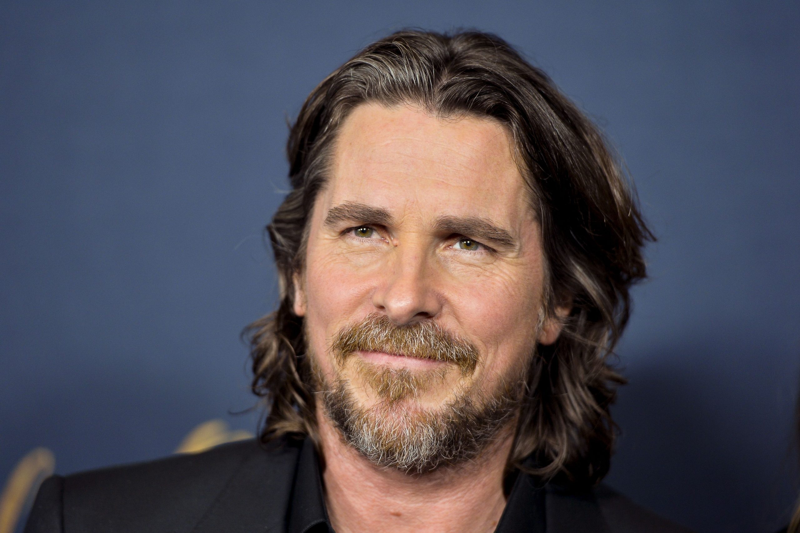Christian Bale smiling warmly on the red carpet at a movie premiere.