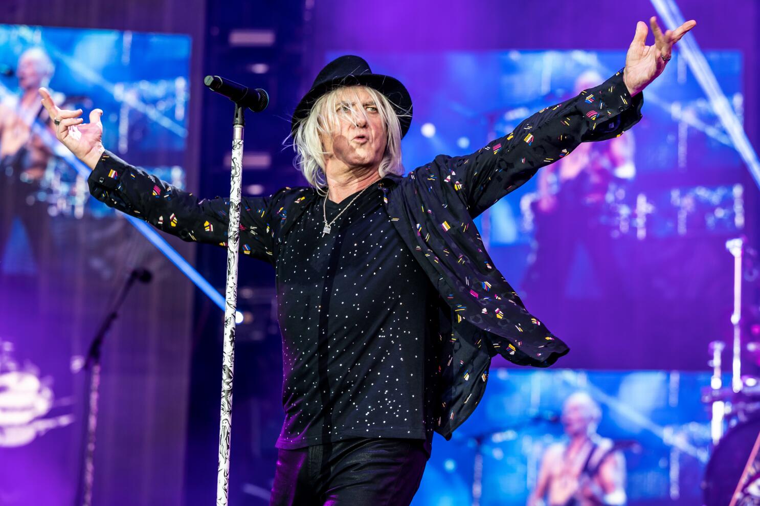 Joe Elliott performing on stage with arms wide open, wearing a stylish fancy hat and engaging the crowd.