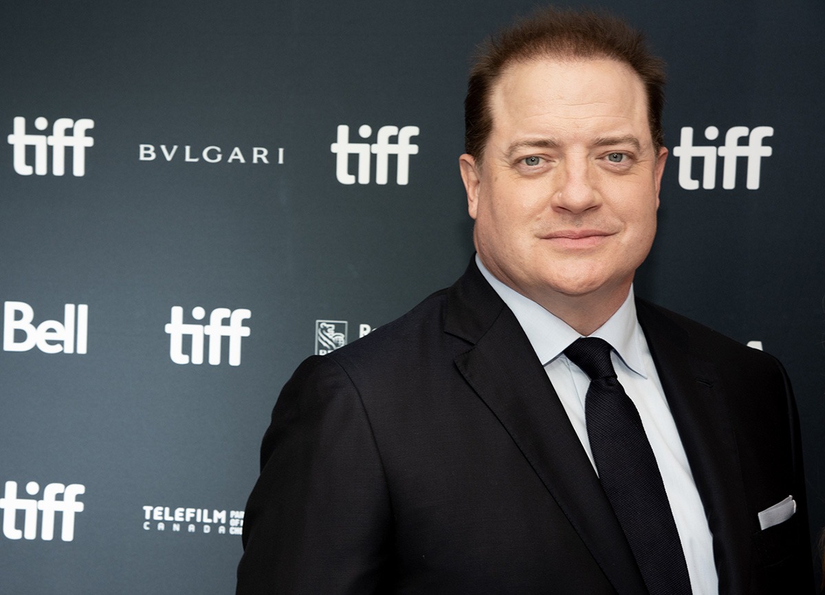 Brendan Fraser wearing a sharp black suit and tie, posing confidently on the red carpet at a premiere event.