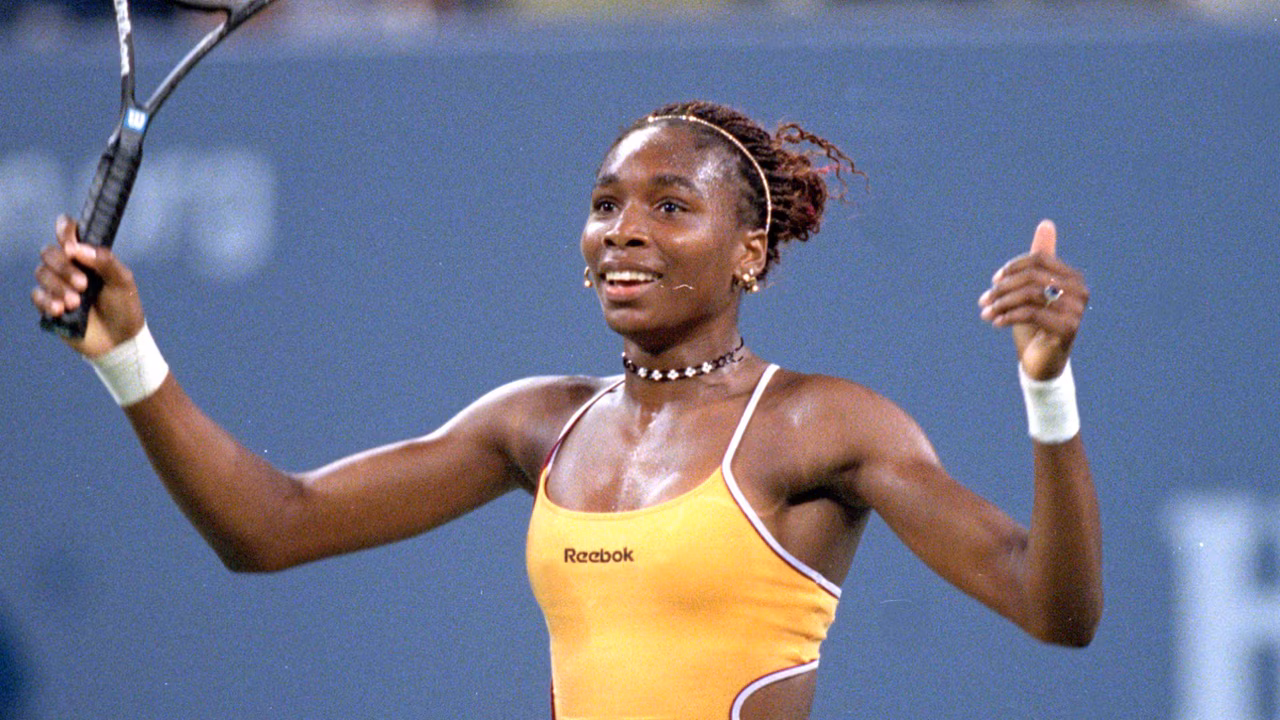 A young Venus Williams smiling brightly, holding her tennis racket triumphantly in the air during her early playing days.