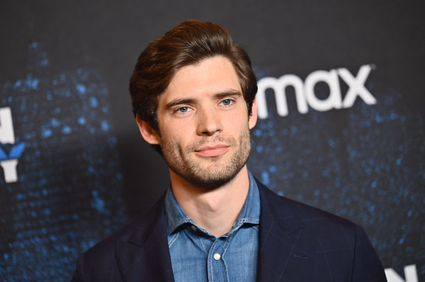 David Corenswet smiling warmly at the camera while posing on the red carpet in a sharp suit.