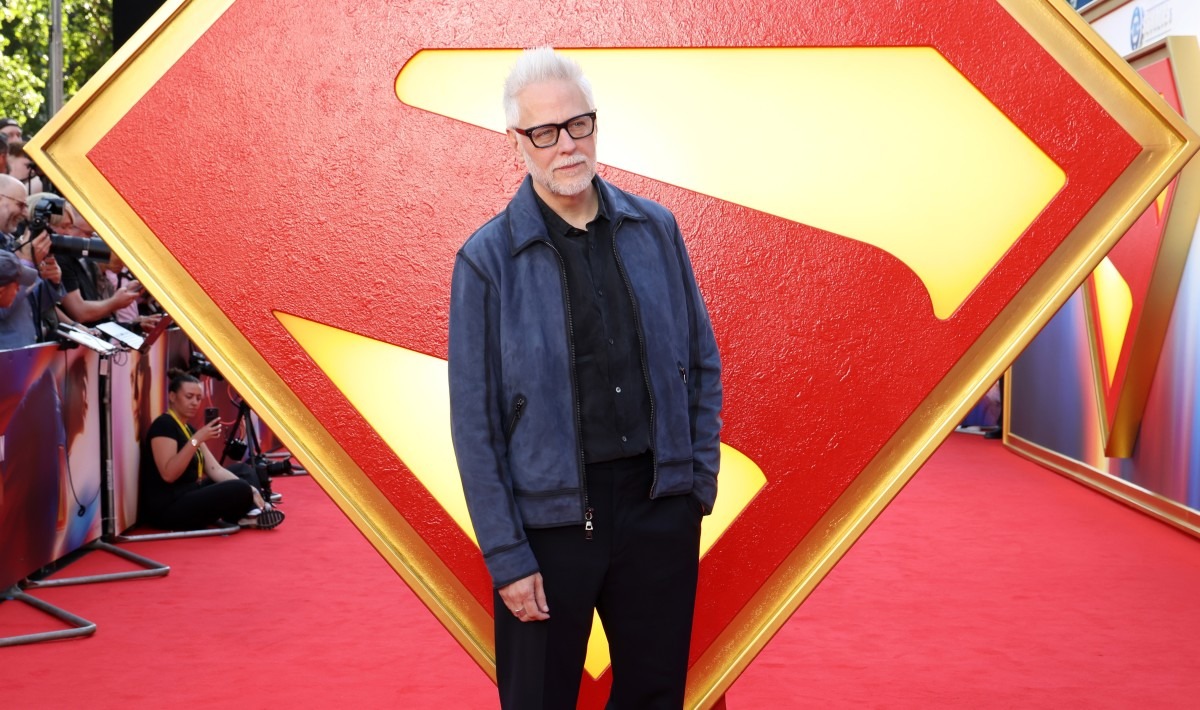 James Gunn standing at the Superman premiere in front of a large Superman logo backdrop.