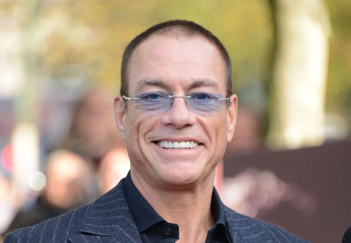 Jean-Claude Van Damme smiling in a suit and blue-tinted glasses at a public event.