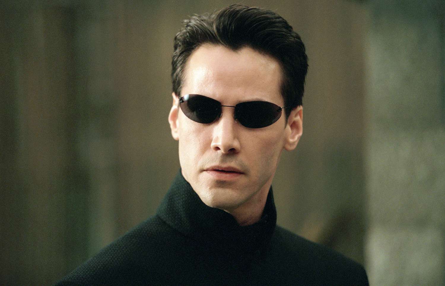 Keanu Reeves as Neo in The Matrix, wearing a black trench coat and sunglasses in a futuristic setting.