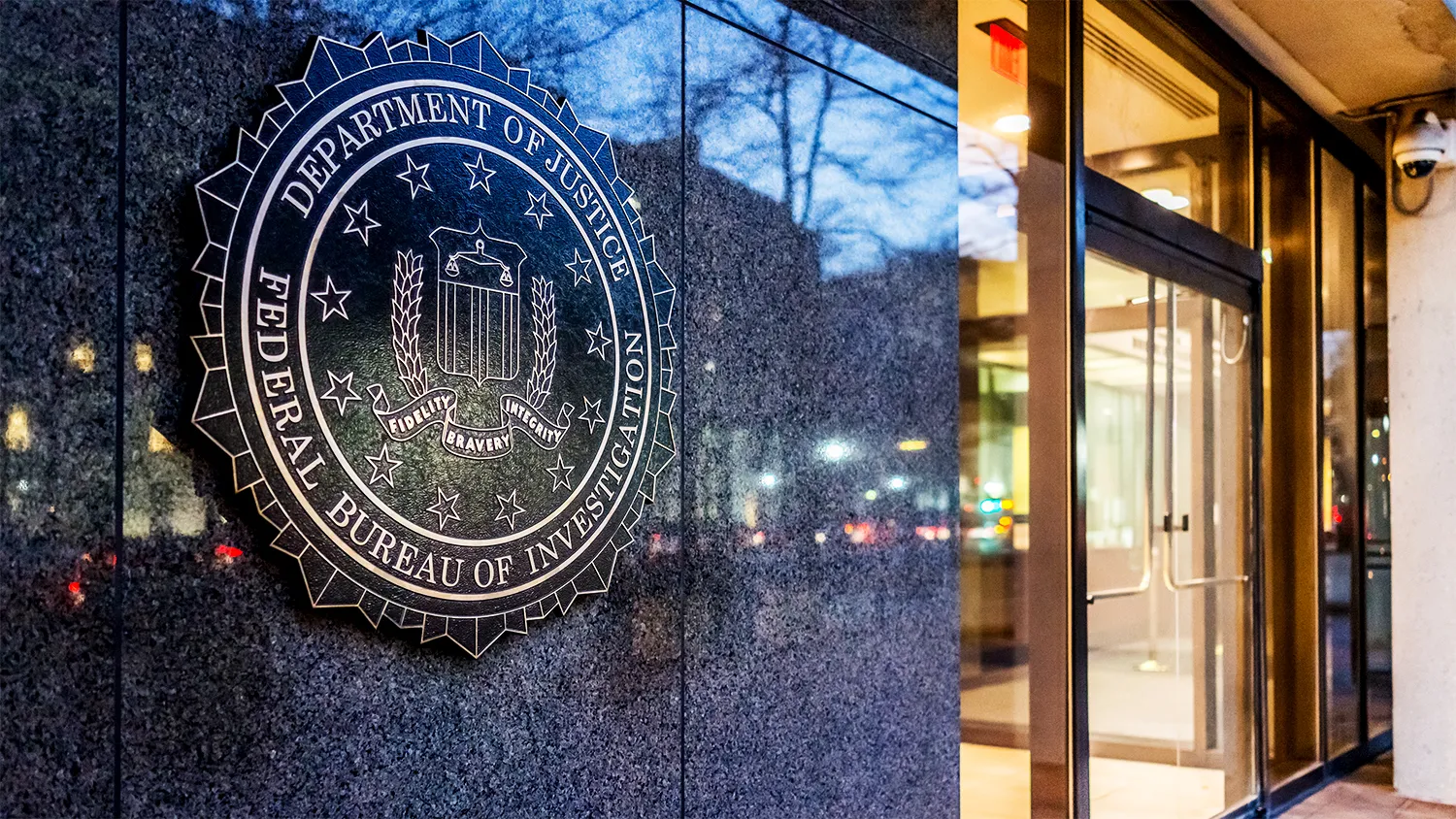 The FBI seal mounted on the granite exterior of the J. Edgar Hoover Building in Washington, D.C., next to the main glass entrance, with reflections of city lights and trees visible in the background.