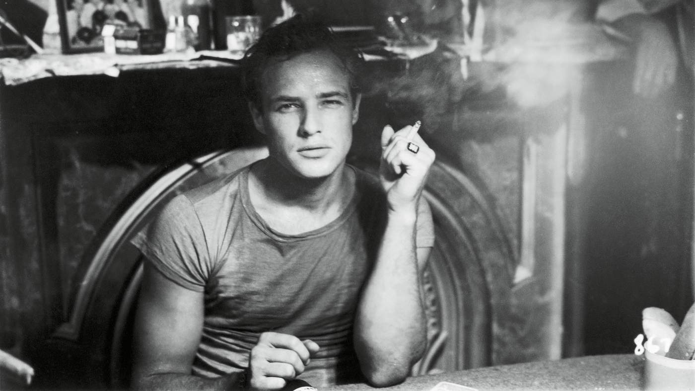 A young Marlon Brando sitting down, smoking, and looking directly into the camera.