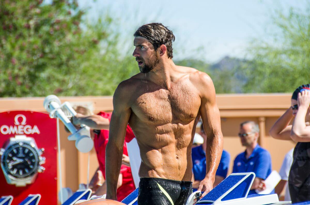 Michael Phelps, visibly muscular and shredded, walking confidently around a poolside.