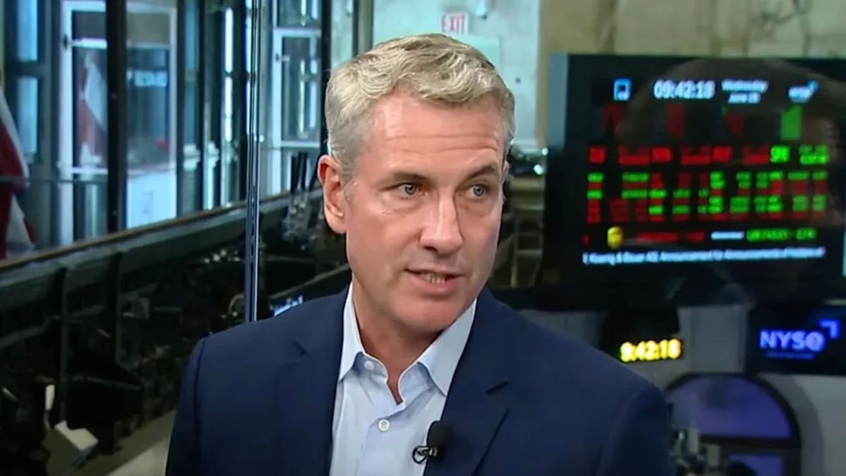 Andy Byron being interviewed with computer screens displaying data behind him.