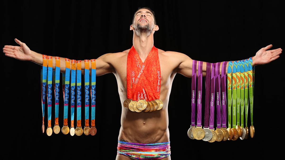 Michael Phelps standing proudly with arms outstretched, his Olympic medals hanging from both arms.