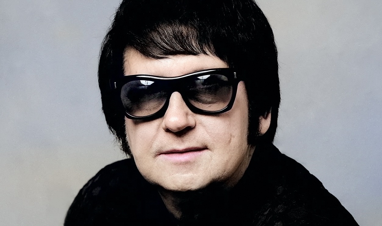 Roy Orbison wearing his signature dark sunglasses and a black outfit, posing against a neutral background.