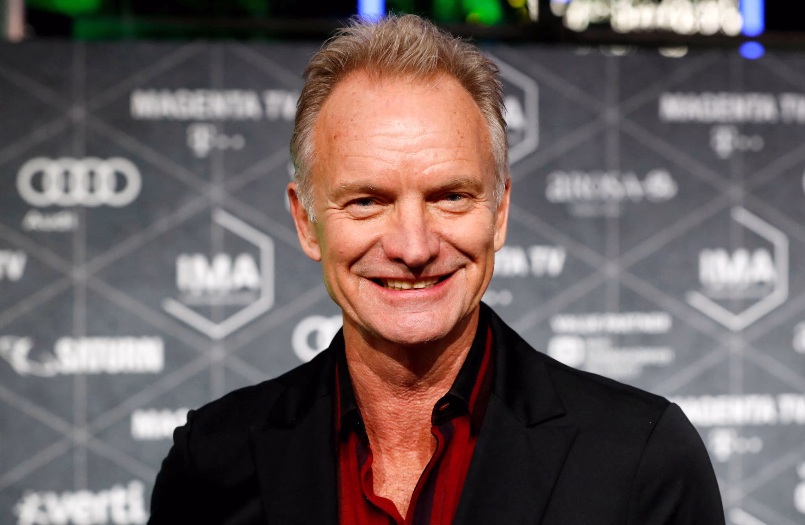 Sting smiles on the red carpet in a black blazer and red shirt at an event, with branded media logos in the background.