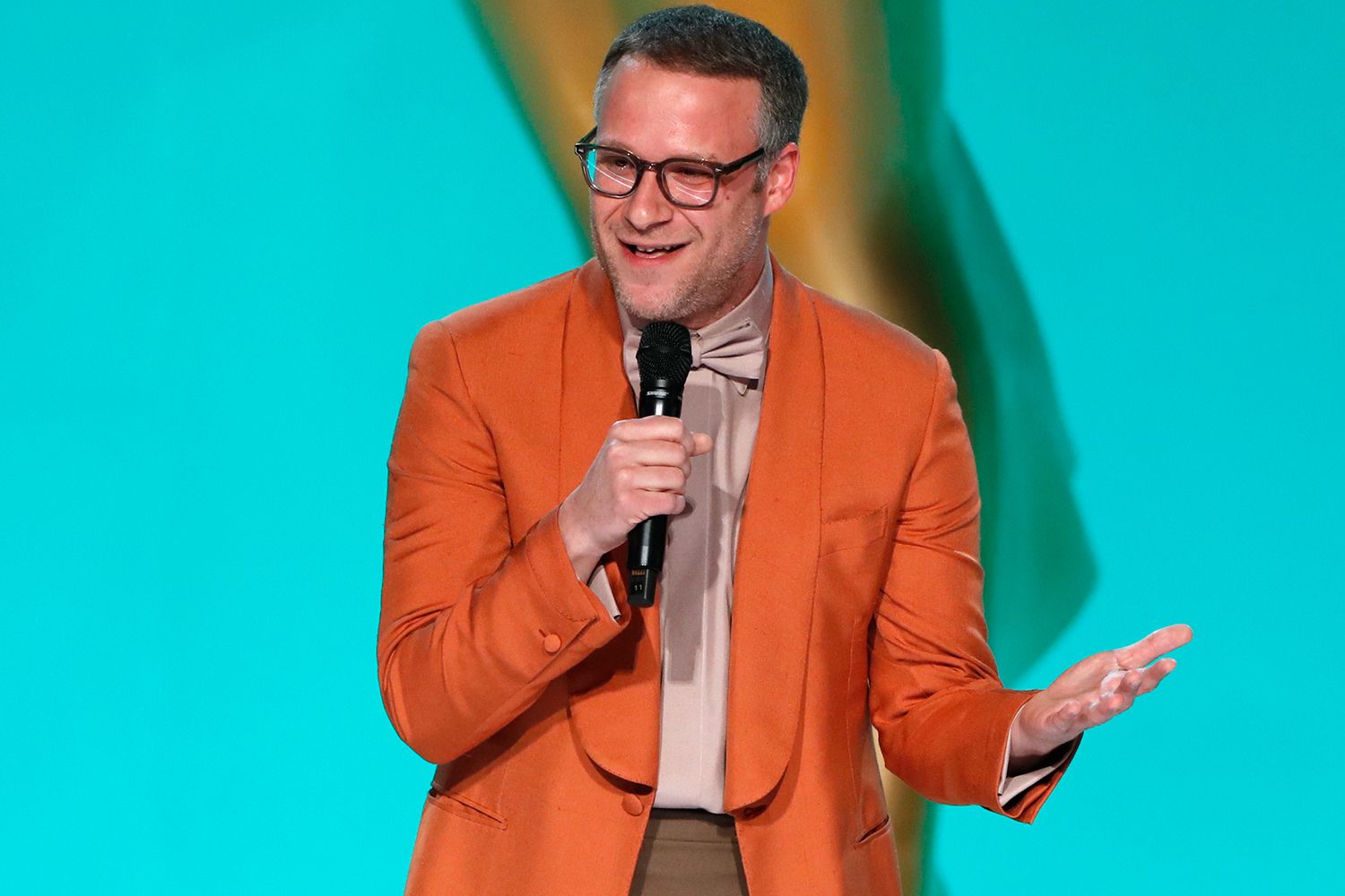 Seth Rogen delivering a speech onstage at the Oscars ceremony, dressed in an Orange blazer.