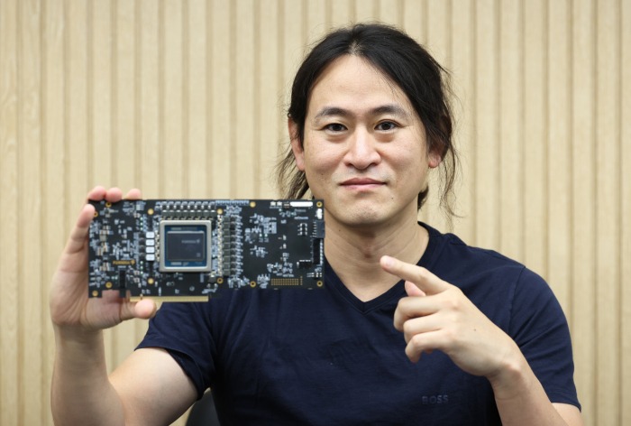 June Paik, co-founder of FuriosaAI, holds and points to the company’s Renegade AI processor board while smiling for the camera.