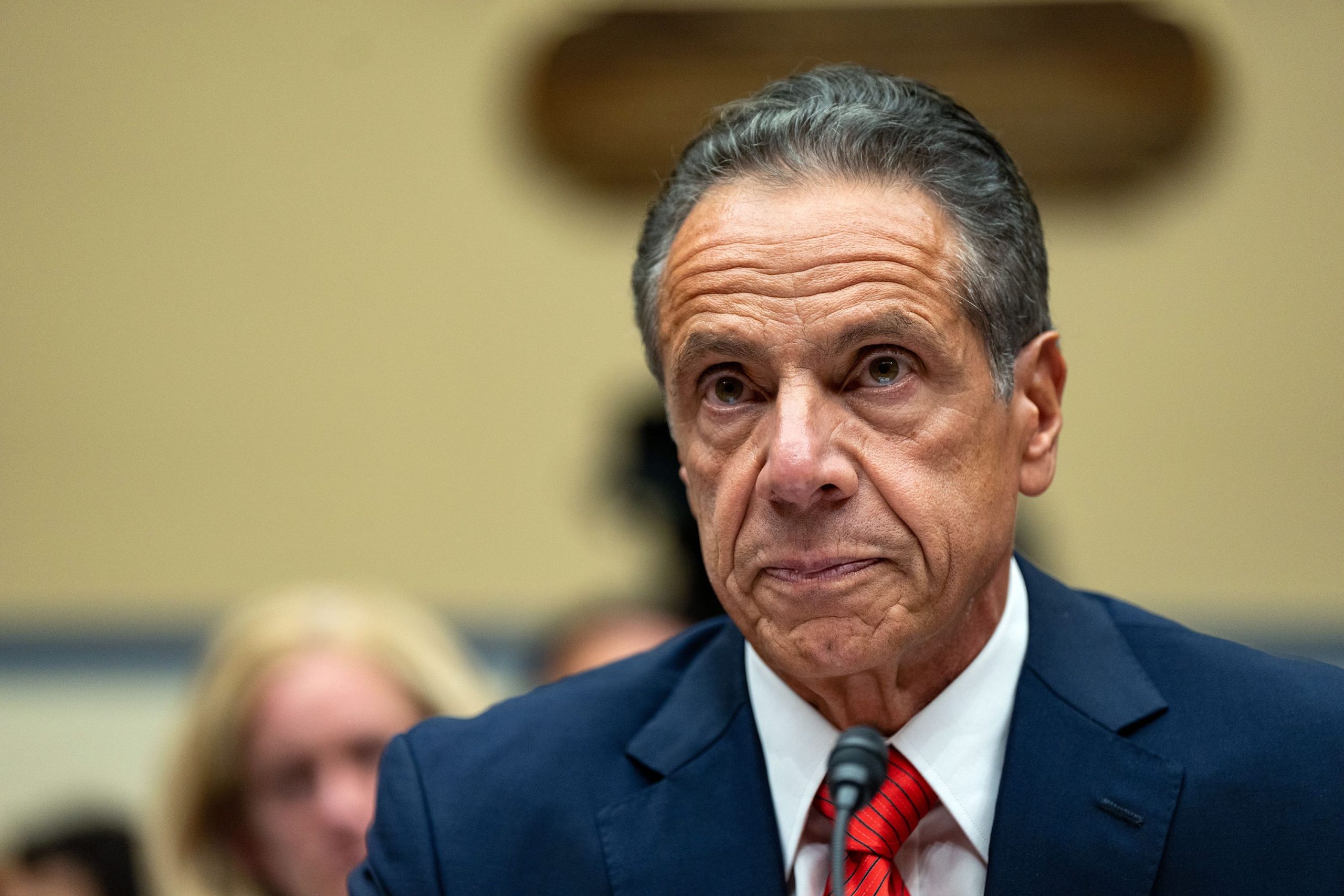 Andrew Cuomo speaks at a public hearing, wearing a navy suit and red tie, appearing serious and focused.