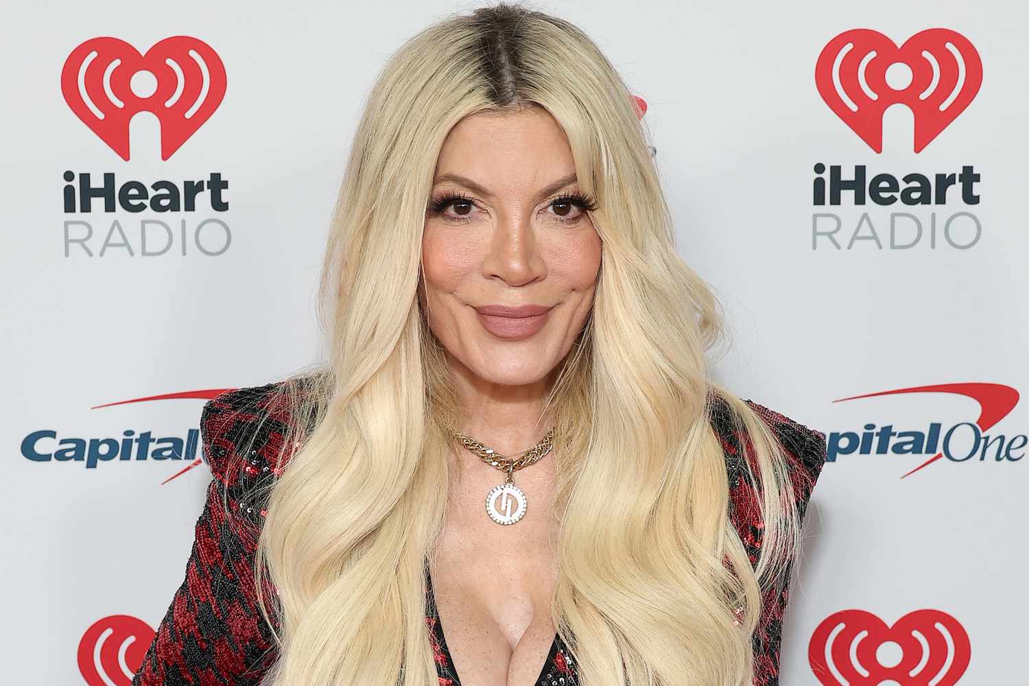 Tori Spelling at the iHeartRadio red carpet event, wearing a red and black outfit with long blonde hair and gold necklace.