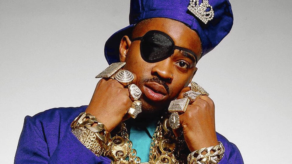 A young Slick Rick dressed in blue, adorned with his signature oversized gold chains, posing confidently for the camera.