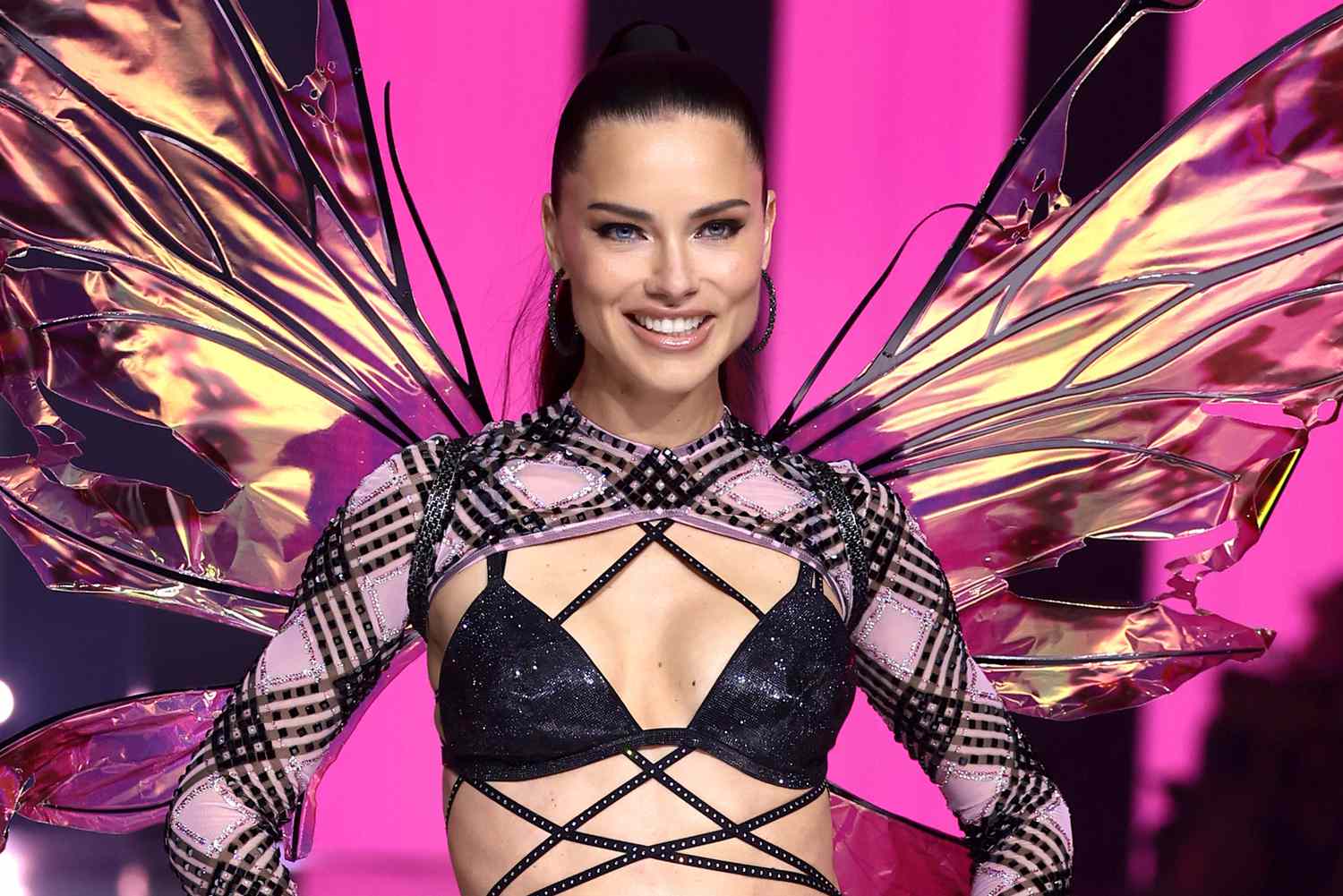 Adriana Lima smiling on the runway in a dazzling costume with wings during a fashion show.