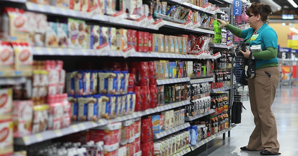 A Walmart employee stocks shelves in the grocery aisle, arranging protein bars and nutritional products in a well-lit store.