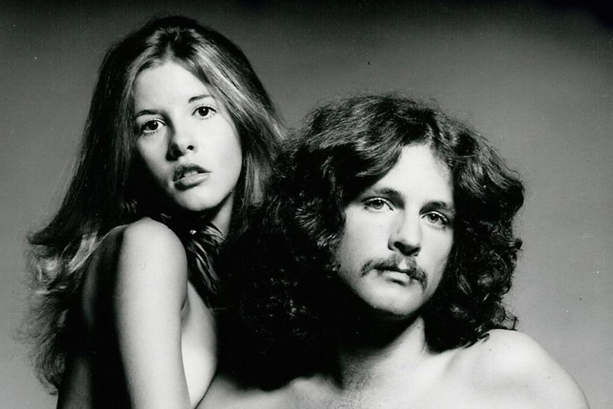 Black and white photo of a young Stevie Nicks and Lindsey Buckingham posing together during a photoshoot.