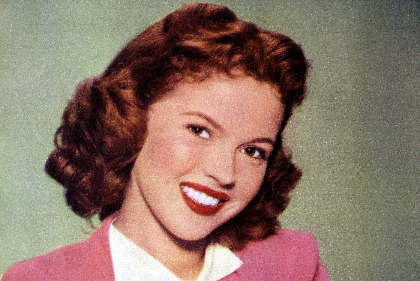 Young Shirley Temple smiling at the camera while wearing a pink dress.